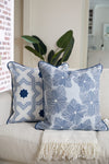 Two blue and white patterned throw pillows on a cream sofa with a fringed blanket add modern flair. One pillow showcases an exclusive Sewing Down South™ geometric star design, while the other is from the Modern Collection: Mod Flora Pillow. A leafy plant and brick wall complete the cozy scene.