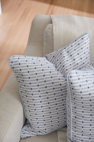 Two Oversized Mod Mini Crossing Tiles in Blue Pillows sit on a light couch beside a textured cream blanket, adding a Southern home decor touch against the backdrop of a wooden floor.