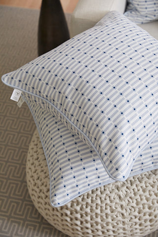 Two Oversized Mod Mini Crossing Tiles in Blue Pillows are stacked on a chunky knit pouf, bringing Southern charm to the room next to a patterned rug and a light-colored sofa.