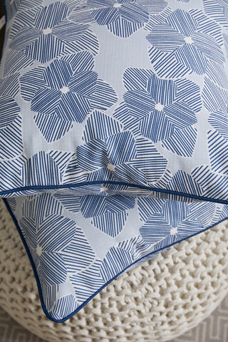 Two Oversized Mod Flora Pillows from Sewing Down South, featuring blue geometric floral patterns on white with blue piped edges and textured fabric, are stacked on a chunky-knit cream pouf.