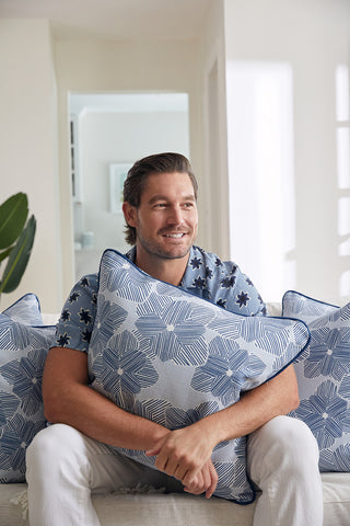 A man with brown hair sits on a white couch, smiling and hugging the Oversized Mod Flora Pillow. Surrounded by other modern decorative pillows, he relaxes in a bright room with natural light and white walls.