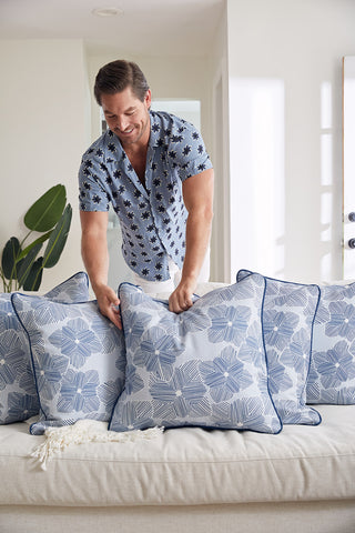 A man in a light blue, short-sleeve floral shirt arranges Sewing Down South’s Oversized Mod Flora Pillow and other modern pillows on a light sofa in a bright, contemporary living room.