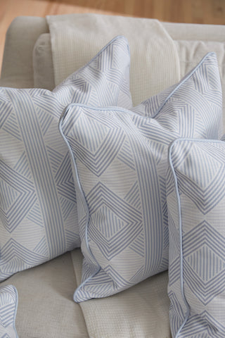 Three Oversized Mod Geo Diamond Pillows in light blue and white geometric patterns rest on a light sofa, complemented by a cream textured blanket. The cozy setting is perfect for enjoying your favorite accent pillow.