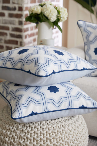 Two Oversized Mod Stella Tile Pillows with blue geometric star patterns are stacked on a chunky knit pouf, with a blurred couch, a matching blue twill pillow, and white flowers in the background.