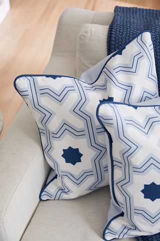 Two Oversized Mod Stella Tile Pillows with blue geometric star patterns rest on a light beige sofa, while a navy knit blanket is draped over the back, creating a bright and cozy scene.