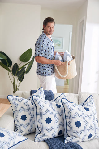 A man smiles while holding a woven basket of decorator pillows, standing in a bright living room with large green plants and a sofa adorned with Oversized Mod Stella Tile Pillows featuring bold geometric designs.