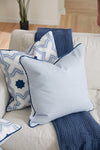 A cozy sofa features a light blue pillow with dark piping from the Sewing Down South Modern Collection: Mod Stella Tile Pillow, paired with a geometric-patterned pillow in blue and white. A textured blue throw blanket is elegantly draped over the armrest.