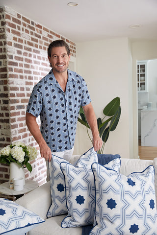 A smiling man in a blue patterned shirt arranges the Oversized Mod Stella Tile Pillow, featuring a geometric design, on a sofa in a bright living room with a brick wall, potted plant, and flowers on the side table.