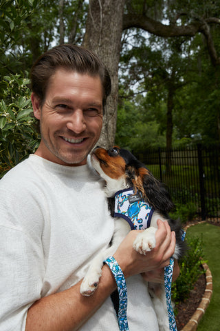 A smiling man in a white shirt holds a small black, brown, and white dog wearing the Crab Craze Dog Harness. The dog nuzzles his face as they pose outdoors with greenery and a black fence in the background.