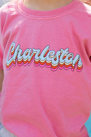 A child in a pink Youth Charleston Tee, made from super soft cotton, stands beside a friend wearing a light blue t-shirt. Charleston is cheerfully printed in colorful, wavy letters on this charming kids top.