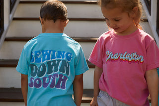 Two young children stand on outdoor steps. The one on the left wears a blue Youth SDS Bubble Tee, while the other sports a pink youth shirt with “Charleston” in colorful text—both are must-have tees for kids.