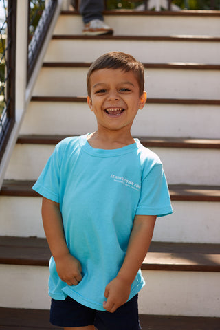 A young boy with short brown hair stands on outdoor wooden stairs, smiling brightly in his Youth SDS Bubble Tee—a light blue shirt—paired with dark shorts and sunlight illuminating his face.
