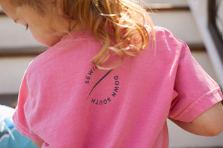 A child with light brown hair stands outdoors by stairs, wearing the stylish Youth Charleston Tee, made of super soft cotton and showcasing the Sewing Down South design with a diagonal line on the back.