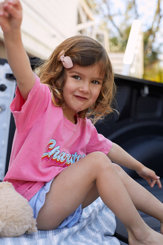 A young girl with wavy brown hair and a pink bow sits in a truck bed, wearing a Youth Charleston Tee and blue shorts. Shes smiling with her arms raised, next to a soft toy. The building and clear sky in the background add charm to the scene.