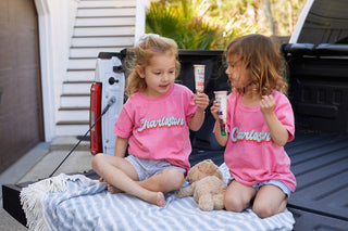 Two young children sit on a blanket in the trucks back, wearing matching Youth Charleston Tees. They enjoy ice cream treats next to a stuffed animal, with stairs and greenery in the background, creating an ideal setting for a delightful afternoon in kids clothing style.