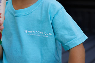 Wearing the Youth SDS Bubble Tee, a light blue shirt with SEWING DOWN SOUTH CHARLESTON, SOUTH CAROLINA on the chest, a child stands holding an object just out of frame to the left. This is the kids favorite tee.