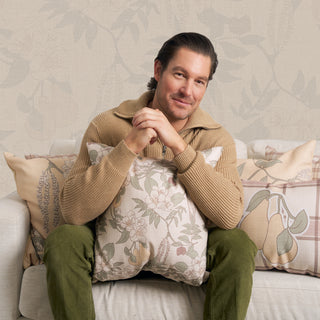 A man with brown hair sits on a sofa, smiling and holding the Fall Florals Pillow. He wears a beige sweater and green pants. Decorative floral pillows line the sofa, set against subtle floral-patterned wallpaper in the background.