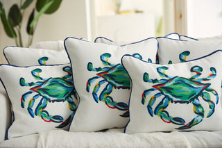 Five pillows from the Coastal Collection, featuring vibrant crabs on white fabric, decorate a couch. Among them is The King Crab Lumbar Pillow with Navy. Crabs are painted in shades of green, blue, and red amidst blurred greenery and light-colored furnishings.