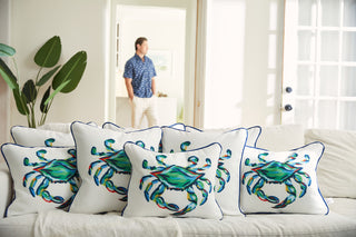 A white couch adorned with The King Crab Lumbar Pillow with Navy and various decorative pillows sits in a bright, sunlit room. A person wearing a blue shirt and white pants stands by the doorway, while a large green plant from the Coastal Collection adds to the ambiance.