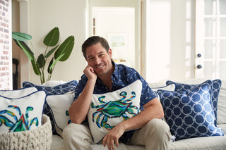A man sits on a sofa, smiling with The King Crab Lumbar Pillow with Navy from the Coastal Collection. He wears a blue patterned shirt and light pants, while a large plant adds to the brightly lit background.