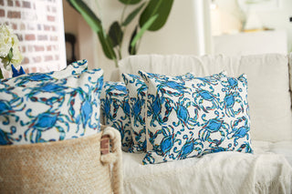 A cozy living room scene features a beige sofa with the Crab Craze Shades of Blue Lumbar Pillow, displaying a charming blue crab design. Verdant leafy plants offer a lush backdrop, and a basket lined with matching Crab Craze fabric anchors the foreground.