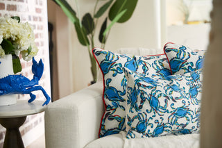 A cozy living room from the Coastal Collection includes a white couch with decorative pillows, featuring the Crab Craze Shades of Blue Lumbar Pillow with red trim. A round table nearby displays a bold blue crab sculpture and a vase of white flowers, set against lush greenery.