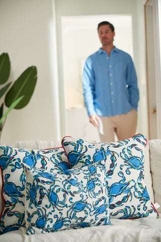 A group of Crab Craze Shades of Blue Lumbar Pillows adorns the sofa with blue crab patterns. A man in a blue shirt and beige pants walks forward, while a large green plant on the left contributes to the serene coastal charm.