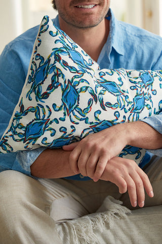 A person in a light blue shirt and beige pants sits, hugging the Crab Craze Shades of Blue Lumbar Pillow, featuring a colorful blue and green leafy pattern. The shot is partially cropped to reveal only their mouth and hands.
