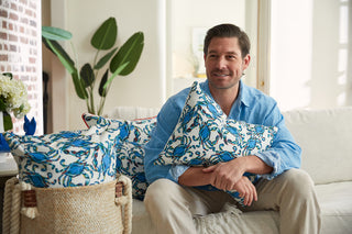 A man in a light blue shirt sits on a white sofa, holding the Crab Craze Shades of Blue Lumbar Pillow. Hes surrounded by coordinating pillows and a woven basket, with a green plant and brick wall in the background, creating a cozy living room atmosphere.