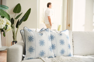 In a bright room, a Coastal Collection sofa is decorated with three Navy Starburst Pillows. A man appears blurred near large windows, where potted plants and white flowers flourish, enhancing the serene ambiance.