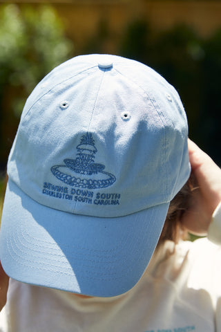 A person wears the Charleston Pineapple Embroidered Hat in light blue, showcasing a pineapple fountain design with Spunkie Down South Charleston, South Carolina text. Against blurred greenery, this premium cotton hat embodies Southern charm with its distinctive motif.