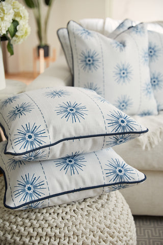 Two Navy Starburst Pillows from the Coastal Collection, featuring dark piping, rest on a knitted surface. More pillows from Sewing Down South can be seen in the background on a cream sofa next to a partially visible potted plant.