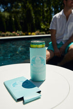 A can in a CHS Pineapple Koozie from the 4 PACK set sits on a round white poolside table, with another drink cooler nearby. In the background, a person in white and teal relaxes by the pool, surrounded by lush greenery.