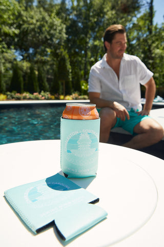 A man in a white shirt and teal shorts sits by a pool. In the foreground, a can with an orange top is in a CHS Pineapple Koozie (4 PACK) on a white table, with another empty drink cooler nearby.
