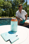 A man in a white shirt and teal shorts sits by a pool. In the foreground, a can with an orange top is in a CHS Pineapple Koozie (4 PACK) on a white table, with another empty drink cooler nearby.