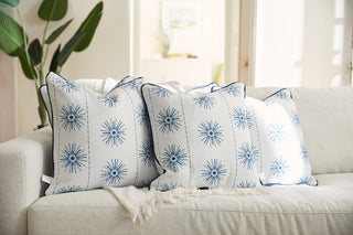 Three Navy Starburst Pillows from the Coastal Collection are placed on a light sofa with a white blanket draped over it. In the background, a plant and blurred wall art add to the cozy setting.