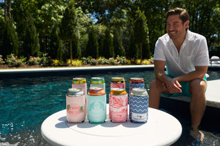 A man in a light shirt and shorts smiles by a pool, with several colorful drinks in neoprene koozies—including a CHS Pineapple Koozie, 4 PACK (Skinny or Standard)—on a round white table. Lush greenery fills the background.