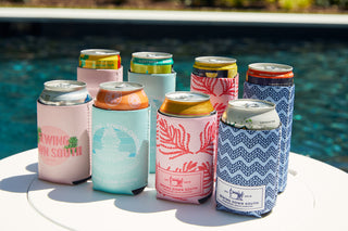 Four CHS Pineapple Koozies (Skinny or Standard) with vibrant South Carolina-inspired designs are arranged on a white table beside a pool, each keeping canned drinks cool in style.