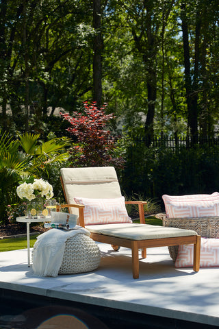 A cushioned wooden lounge chair with a Coral Greek Key Extended Lumbar Outdoor Pillow sits on a patio in a lush garden, next to a white side table holding flowers, drinks, and a basket. Dappled sunlight filters through the trees behind.