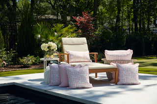 A wooden lounge chair with beige SDS Shield® Outdoor Fabric cushions and a Coral Greek Key Extended Lumbar Outdoor Pillow sits on a patio near a garden, next to a side table with flowers and drinks, surrounded by lush greenery and trees.