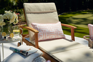 A cushioned lounge chair with a Coral Greek Key Extended Lumbar Outdoor Pillow sits by a small round table holding lemon water, glasses, and white flowers. Nearby are a magazine and blanket, all set on a green lawn—perfect for relaxing outdoors.