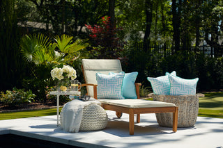 A wooden outdoor lounge chair with blue and white pillows, featuring the Mint Greek Key Outdoor Pillow, sits by a small table with flowers and lemonade. A knitted pouf and a basket of pillows complete the lush garden setting.