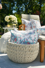 In a cozy outdoor seating area, a woven basket displays Coral Crab Craze Outdoor Pillows. A wooden lounge chair featuring a Sewing Down South™ cushion sits nearby, alongside a small table with flowers and lemonade glasses, all framed by lush greenery.
