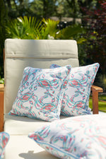 The outdoor patio chair, adorned with two colorful pattern pillows like the Coral Crab Craze Outdoor Pillow, showcases a vibrant mix of pink, blue, and green hues. Surrounded by lush greenery and sunlit leaves, it crafts a peaceful and welcoming ambiance.