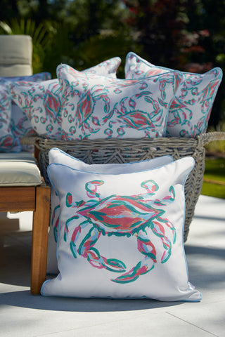 The Coral Crab Craze Lumbar Outdoor Pillow, boasting vivid crab designs and a Caroline Blue welt on SDS Shield® Outdoor Fabric, adds flair to a woven basket and wooden bench in a lush green outdoor setting under the sun.