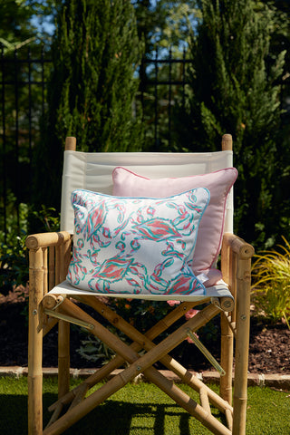 A bamboo-framed chair with a cream seat is decorated with the colorful Coral Crab Craze Lumbar Outdoor Pillow. The outdoor scene, enhanced by green trees and shrubs, offers a tranquil and stylish atmosphere.
