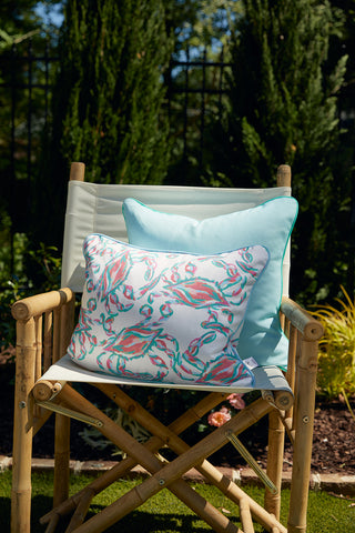 A bamboo chair with a white cushion holds two pillows: one solid light blue and one Mint Greek Key Outdoor Pillow. The chair is outdoors, surrounded by greenery and sunlight.