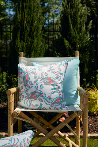 A wooden directors chair with light fabric is outdoors, featuring the Coral Crab Craze Outdoor Pillow in the center. A smaller blue cushion is alongside it, framed by lush greenery and a black fence in the background.