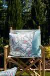 A wooden directors chair with light fabric is outdoors, featuring the Coral Crab Craze Outdoor Pillow in the center. A smaller blue cushion is alongside it, framed by lush greenery and a black fence in the background.
