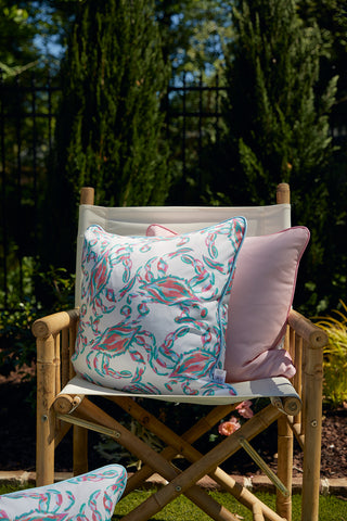 A decorative chair with the Coral Crab Craze Outdoor Pillow featuring a Sewing Down South™ design in pink and green florals is on a sunny patio. Behind the chair, lush shrubs and a black metal fence enhance the inviting garden backdrop.